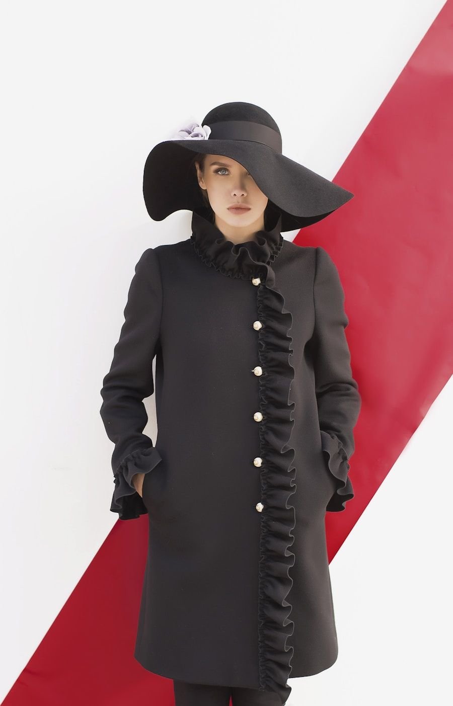 Model in a long black silk coat and wide-brimmed hat against a graphic red and white backdrop